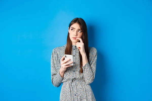 Chica linda pensativa pensando cómo responder en el mensaje, mirando a un lado reflexivo y sosteniendo el teléfono inteligente, de pie en vestido sobre fondo azul —  Fotos de Stock