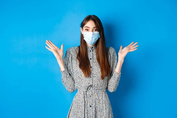 Pessoas saudáveis e conceito de pandemia covid-19. Menina surpresa em máscara médica levantando as mãos para cima animado, ouvir boas notícias, sorrindo com os olhos, de pé fundo azul — Fotografia de Stock