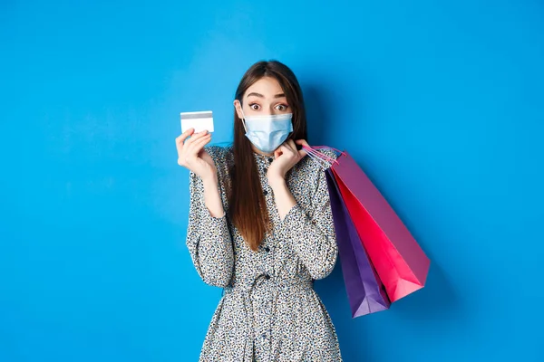 Covid-19, concept de pandémie et mode de vie. Femme excitée portant un masque médical lors de vos achats, montrant une carte de crédit en plastique et des sacs de retenue, fond bleu — Photo