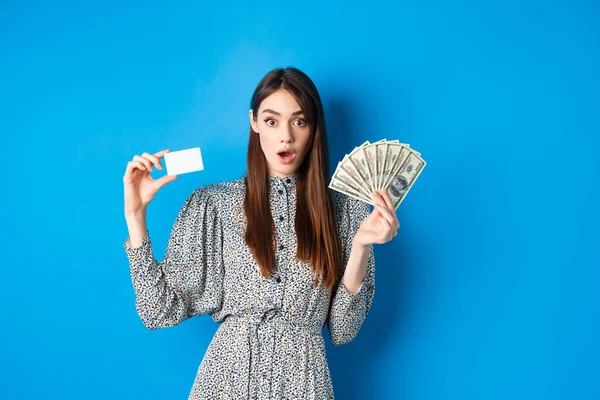 Muchacha afortunada emocionada mostrando tarjeta de crédito de plástico y dinero dólar, jadeando sorprendido, comprando algo caro, de pie sobre fondo azul — Foto de Stock