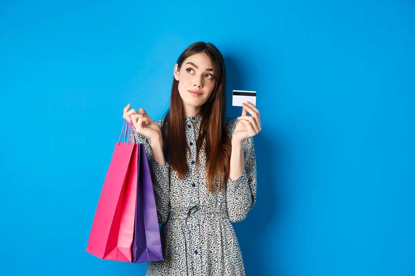 Zasněný dívka dívá na logo a ukazuje plastové kreditní karty, držení nákupních tašek, stojící na modrém pozadí — Stock fotografie