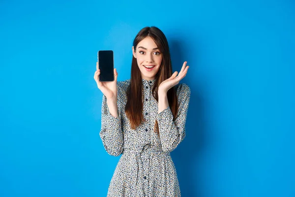 Возбужденная девушка показывает пустой экран смартфона и задыхаясь очарован, продемонстрировать торговый приложение, стоя на синем фоне — стоковое фото