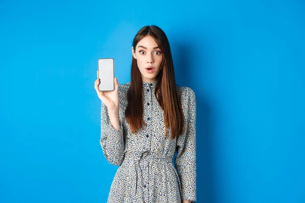 Mujer joven emocionada en vestido, jadeando sorprendido, mostrando la pantalla vacía del teléfono inteligente y mirar impresionado a la cámara, fondo azul —  Fotos de Stock