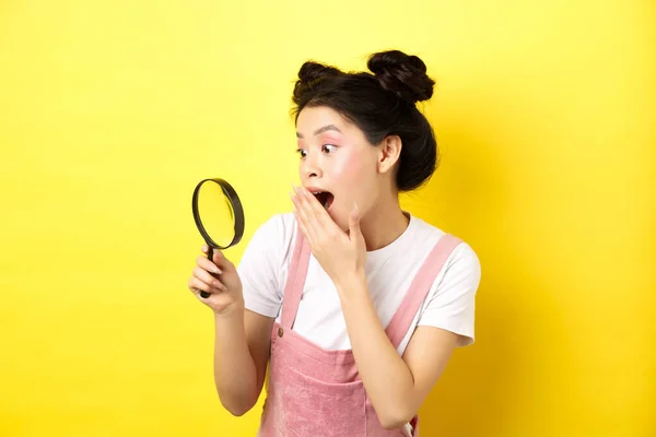 Podekscytowana dziewczyna azjatycka dysząca zastanawiała się, znalazła coś ciekawego, patrząc przez lupę, stojąc na żółtym tle — Zdjęcie stockowe