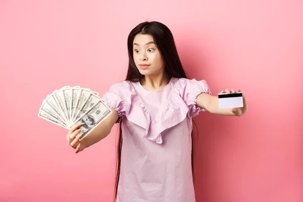 Spännande tonåring asiatisk flicka sträcka ut händer med dollarsedlar och plast kreditkort, titta på pengar med förvåning, står på rosa bakgrund — Stockfoto