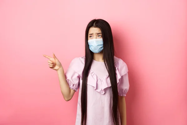 Coronavirus, Quarantäne und Lebensstilkonzept. Traurige und düstere asiatische Frau in medizinischer Maske, die aufgebracht nach links schaut, unfaire Dinge zeigt, auf rosa Hintergrund stehend — Stockfoto