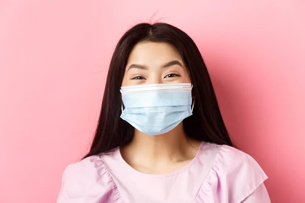 Covid-19 και υγιείς άνθρωποι έννοια. Κοντινό πλάνο του χαρούμενου κοριτσιού από την Ασία που φορούσε ιατρική μάσκα και χαμογελούσε με τα μάτια, στεκόταν πάνω σε ροζ φόντο — Φωτογραφία Αρχείου