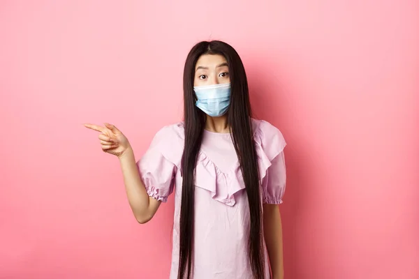 Ковид-19, концепция пандемического образа жизни. Любопытная азиатка в медицинской маске, указывающая налево на логотип, задающая вопросы о повышении, стоящая на розовом фоне — стоковое фото