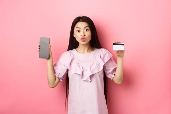 Интернет-магазины. Возбужденная азиатка показывает пластиковую кредитную карту с пустым экраном смартфона, рекламный интернет-магазин, стоя на розовом фоне — стоковое фото