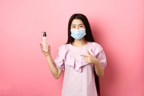 Sağlıklı insanlar ve covid-19 salgını. Tıbbi maskeli neşeli Asyalı kadın el dezenfektanı öneriyor, antiseptik şişeyi işaret ediyor, beyaz arka plan — Stok fotoğraf