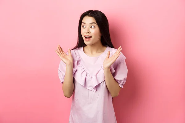 Emocionado chica asiática aplauden las manos y mirando a la izquierda, viendo la actuación y aplausos, de pie en vestido sobre fondo rosa — Foto de Stock
