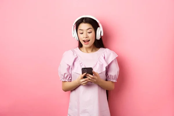 Szczęśliwa piękna kobieta w słuchawkach bezprzewodowych oglądanie wideo na smartfonie, patrząc na zabawę telefonu, stojąc na różowym tle — Zdjęcie stockowe