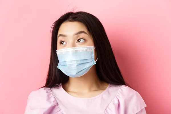 Covid-19, conceito de pandemia e quarentena. Close-up de menina asiática sonhadora em máscara médica olhando à esquerda no logotipo com rosto pensativo, de pé sobre fundo rosa — Fotografia de Stock