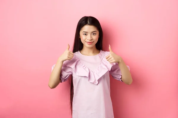 Muito bem. Sorrindo menina asiática no vestido mostrando polegares para cima, como e aprovar o produto, elogiar muito, de pé contra fundo rosa — Fotografia de Stock