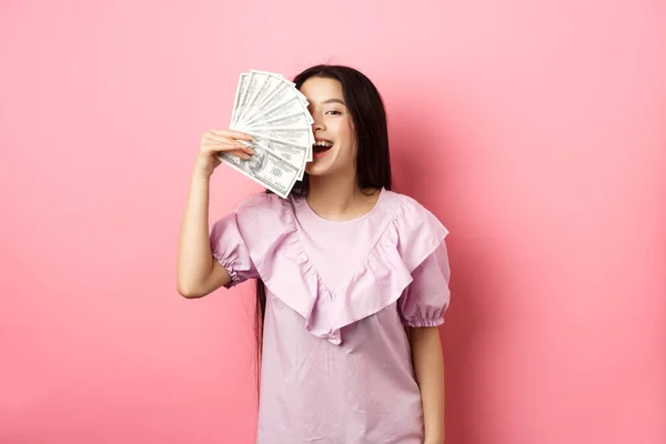 Feliz rico asiático mujer mostrando dinero y sonriendo, compras con efectivo, de pie en vestido sobre rosa fondo — Foto de Stock