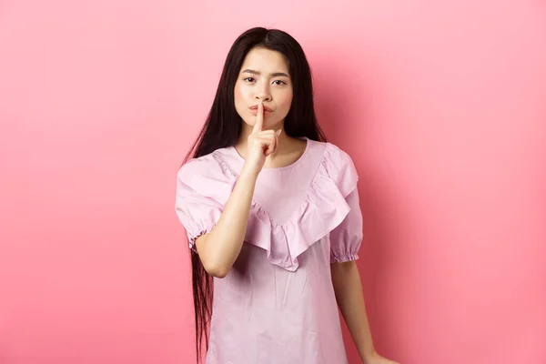 Χαριτωμένο κορίτσι της Ασίας λένε να είναι ήσυχο, κατσαδιάζοντας δυνατά πρόσωπο, που δείχνει σιωπή σημάδι με το δάχτυλο πιέζεται στα χείλη, στέκεται στο φόρεμα σε ροζ φόντο — Φωτογραφία Αρχείου