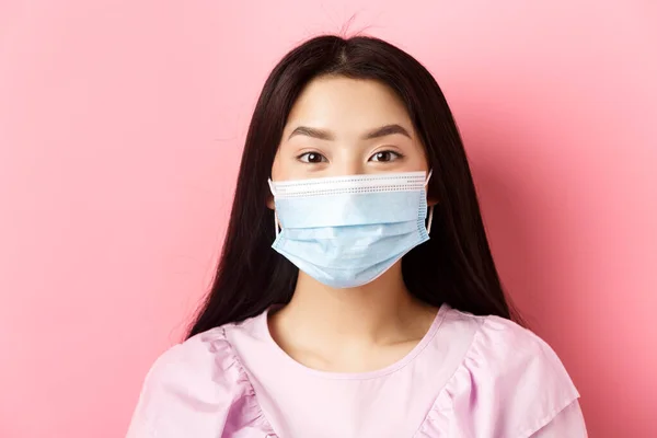Covid-19 και υγιείς άνθρωποι έννοια. Κοντινό πλάνο του ασιατικού χαριτωμένο κορίτσι φορώντας ιατρική μάσκα κατά τη διάρκεια της επιδημίας του ιού, στέκεται πάνω σε ροζ φόντο — Φωτογραφία Αρχείου