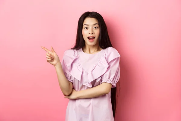 Excitada bela menina adolescente asiática apontando dedo esquerdo, boca aberta espantada e olhando para a câmera, mostrando negócio promo legal, fundo rosa — Fotografia de Stock