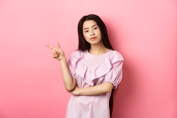 Giovane compiaciuta ragazza asiatica cercando fresco e puntare il dito sinistro al logo, prodotto pubblicitario su sfondo romantico rosa — Foto Stock