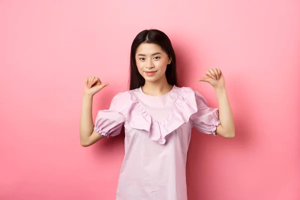 Αυτοπεποίθηση Ασιάτισσα χαμογελά, δείχνει τον εαυτό της, αυτοπροωθείται προσωπικά επιτεύγματα, στέκεται ντυμένη σε ροζ φόντο — Φωτογραφία Αρχείου