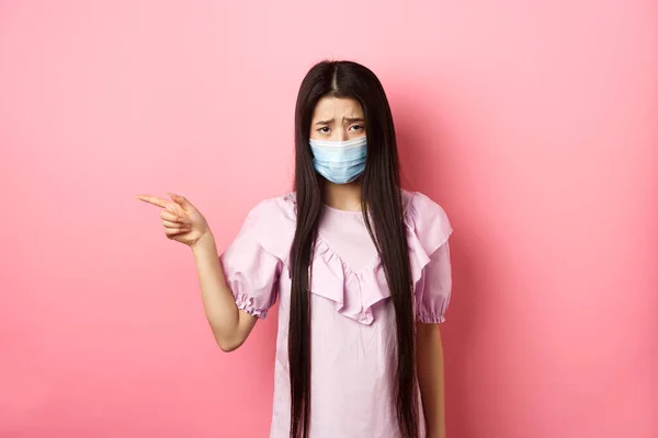 Covid-19, konsep gaya hidup pandemi. Gadis asia yang sedih dan suram dalam topeng medis menunjuk ke kiri, mengeluh tentang situasi yang tidak adil, berdiri kecewa terhadap latar belakang merah muda — Stok Foto