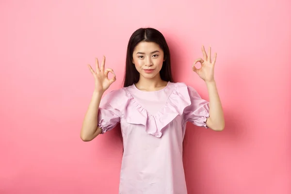 Sonriendo mujer asiática mostrando signos de bien y mirando confiado, asegurar todo lo bueno, alabanza buen trabajo, buen gesto de elección, de pie sobre fondo rosa —  Fotos de Stock