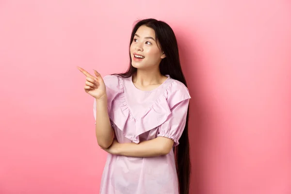 Ενθουσιασμένη χαμογελαστή Ασιάτισσα με φόρεμα, δείχνει και κοιτάζει αριστερά στο λογότυπο, τσεκάρει την προαγωγή, στέκεται σε ροζ φόντο — Φωτογραφία Αρχείου