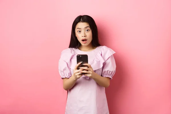 Handla på nätet. Förvånad asiatisk kvinna i söt klänning, öppen mun förvånad efter att ha läst nyheter på smartphone, står på rosa bakgrund — Stockfoto