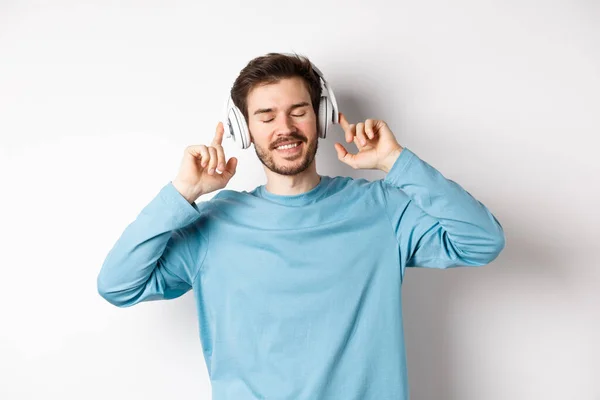 Bonito homem ouvindo música em fones de ouvido sem fio e sorrindo, desfrutando de bom som, fundo branco — Fotografia de Stock