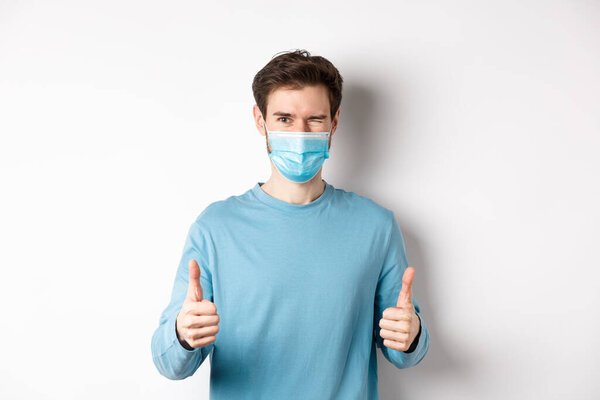 Covid-19, pandemic and social distancing concept. Happy young man in medical mask winking, showing thumbs up in approval, recommending product, white background