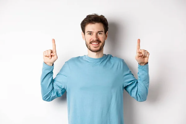 Beeld van de jonge moderne man met baard tonen reclame op kopieerruimte, wijzen vingers omhoog en glimlachen, check it out gebaar, witte achtergrond — Stockfoto