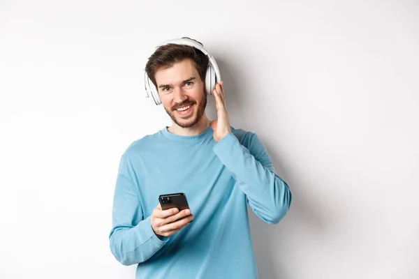 Bonito cara ouvir música em fones de ouvido sem fio, sorrindo para a câmera e usando o smartphone, de pé sobre fundo branco — Fotografia de Stock