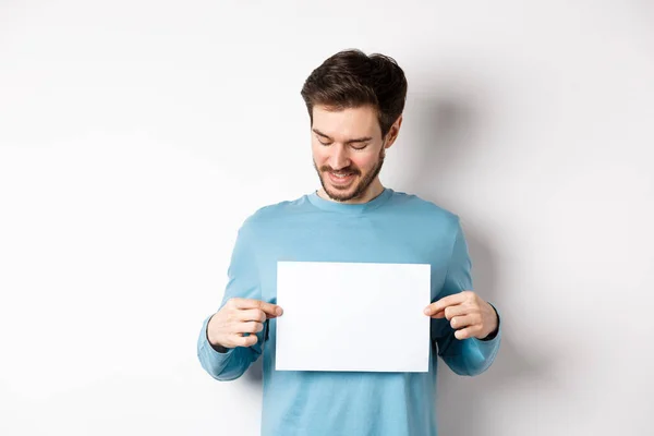 Joven hombre caucásico mirando papel vacío, leyendo el logo, de pie sobre fondo blanco — Foto de Stock