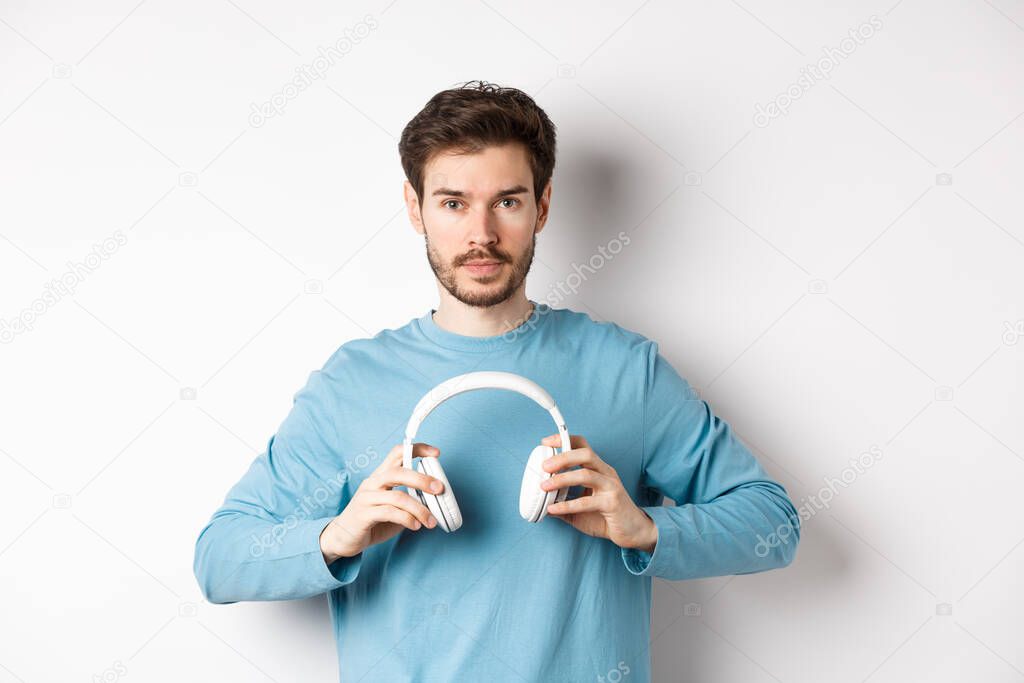 Young bearded guy in blue sweatshirt put on wireless headphones, listening music, standing on white background