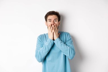 Şok olmuş ve ürkmüş, nefes nefese kalmış, ağzı ellerle kaplanmış, korkunç bir şeye bakıyor, beyaz arka planda duruyor.