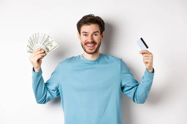Chico feliz sonriendo y mostrando tarjeta de crédito de plástico con dinero en efectivo, elegir entre el dinero y los pagos sin contacto, fondo blanco — Foto de Stock