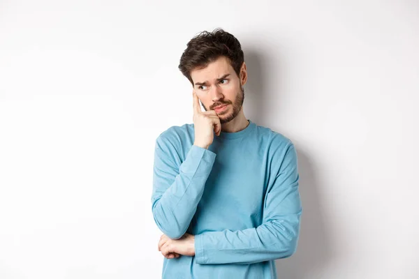 Worried young man with beard in sweatshirt, looking away pensive and thinking, standing troubled against white background — Stock Photo, Image