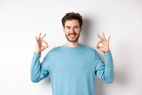 ¡Muy bien! Sonriente chico guapo mostrando signos de bien y buscando satisfecho, de pie sobre fondo blanco, aprobar excelente opción — Foto de Stock