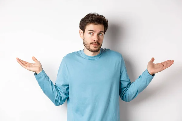 Jeune homme indécis à la barbe, haussant les épaules et ne sachant rien, détournant les yeux confus, debout en chemise bleue sur fond blanc — Photo