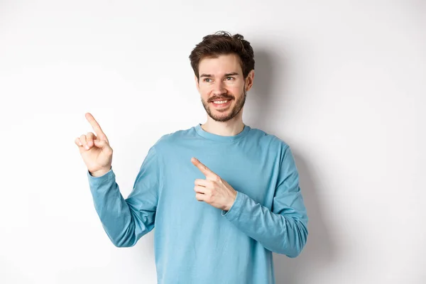 Portrait of young modern man in blue sweatshirt pointing fingers at upper left corner and looking away, smiling pleased as showing advertisement on white background — Stock Photo, Image