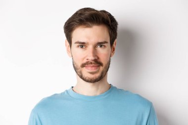 Sakallı, kameraya özgüvenle gülümseyen, beyaz arka planda duran genç ve yakışıklı adamı yakından çek.