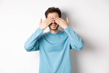 Gülümseyen beyaz adam sürpriz bekliyor, gözleri kapalı, hediye bekliyor, beyaz arka planda duruyor. Kutlama ve tatil kavramı