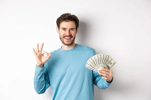 Hombre joven guapo mostrando préstamos rápidos dinero, hacer buen gesto y sonriendo con dinero en efectivo, de pie sobre fondo blanco —  Fotos de Stock