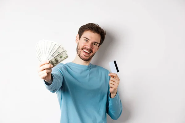Hombre joven guapo en camisa azul que le da dinero, la celebración de la tarjeta de crédito de plástico, de pie sobre fondo blanco — Foto de Stock