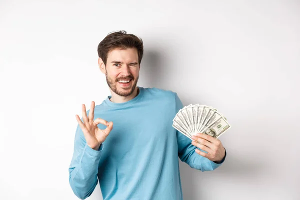 Bezczelny uśmiechnięty mężczyzna mruga, pokazując Ok znak i trzymając pieniądze, koncepcja szybkiego kredytu lub kredytu, stojąc na białym tle — Zdjęcie stockowe