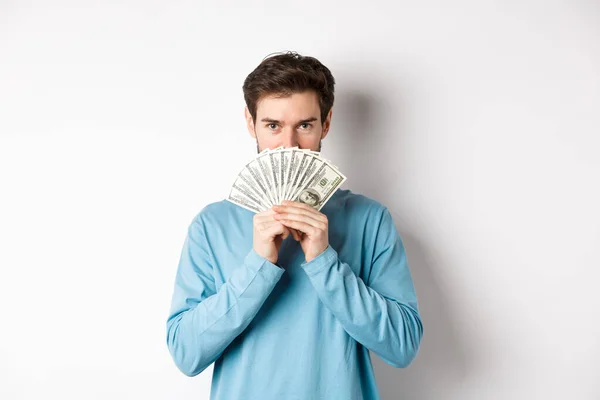 Concepto financiero. Sonriente joven cubriendo la cara detrás del dinero, mostrando dólares y luciendo feliz, de pie sobre fondo blanco — Foto de Stock