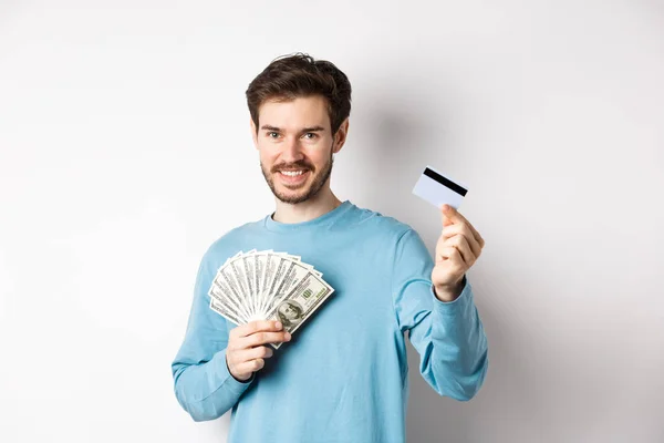 Joven guapo sonriendo y ofreciendo pago en efectivo y sin contacto, mostrando dinero con tarjeta de crédito de plástico, de pie sobre fondo blanco — Foto de Stock