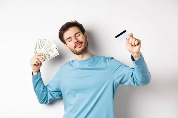 Modelo masculino despreocupado bailando con dinero y tarjeta de crédito, sonriendo y soñando con ir de compras, de pie sobre fondo blanco — Foto de Stock