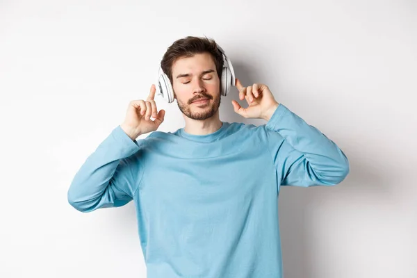 Avslappnad ung man njuter av favoritsång, lyssna musik i hörlurar med slutna ögon och lugnt ansikte, står över vit bakgrund — Stockfoto