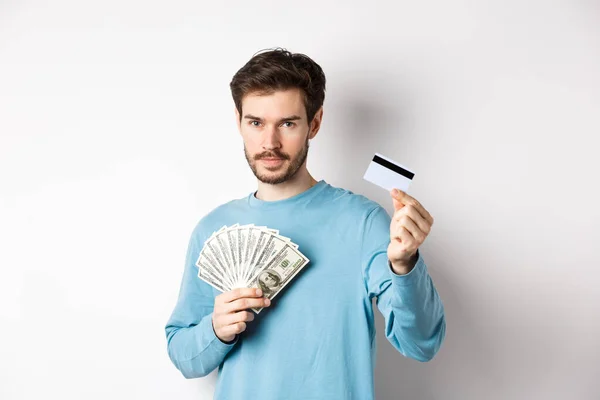 Hombre caucásico sosteniendo dinero y mostrando tarjeta de crédito de plástico, de pie en sudadera azul sobre fondo blanco — Foto de Stock